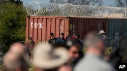 Members of Congress, bottom, look around as migrants walk near a rail car covered in concertina wire at the Texas-Mexico border, in Eagle Pass, Texas, Jan. 3, 2024.
