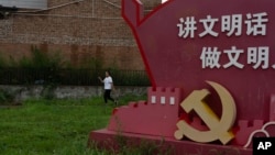 FILE - A woman passes near a government propaganda with the words "Speak in civilized manners, act in civilized manners" near the emblem for the Communist Party on the outskirts of Beijing, July 25, 2024.